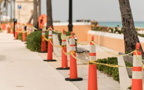 Orange cones and yellow caution tape surround some palm trees on a sidewalk area under construction.