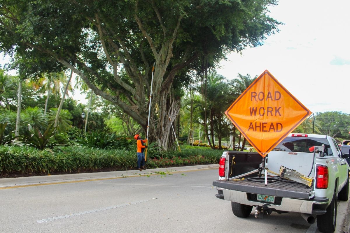 An orange sign reads "Road Work Ahead" as Sherlock Tree Company employees use stick pruners to prune trees in the background.