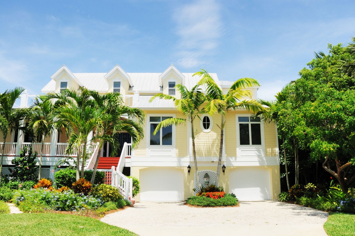 Yellow two-story South Florida home with white metal roof surrounded by healthy palm trees and tropical landscaping on a sunny day.